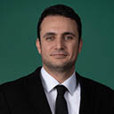 Pessoa adulta com cabelo curto e escuro vestindo terno preto, camisa branca e gravata preta, posando com fundo verde. Yapay zeka taraf ndan olu turulan a  klama